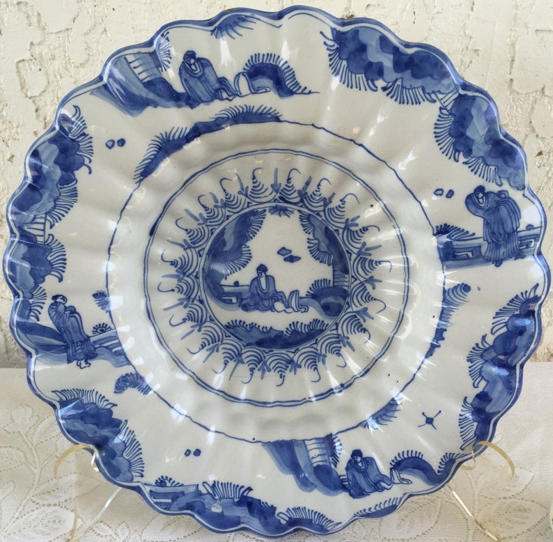 K&ouml;nnte beinhalten: Ein gro&szlig;er, wei&szlig;er Teller aus Keramik mit einem blauen und wei&szlig;en Design. Der Teller hat einen gewellten Rand und zeigt ein zentrales Design einer Figur in einer Landschaft. Der Teller ist mit stilisierten B&auml;umen und Blumen verziert.