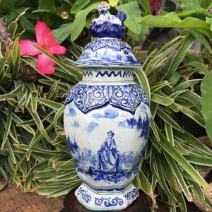 May include: A blue and white ceramic vase with a lid, featuring detailed landscape and figure paintings. The vase has a bulbous shape and stands on a dark wooden base. The lid is topped with a small decorative figure. The vase is surrounded by green plants.