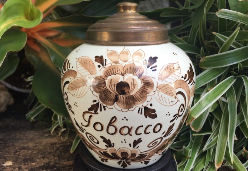 May include: A white ceramic tobacco jar with a brown floral design and a brass lid. The jar has the word "Tobacco" written on the front.