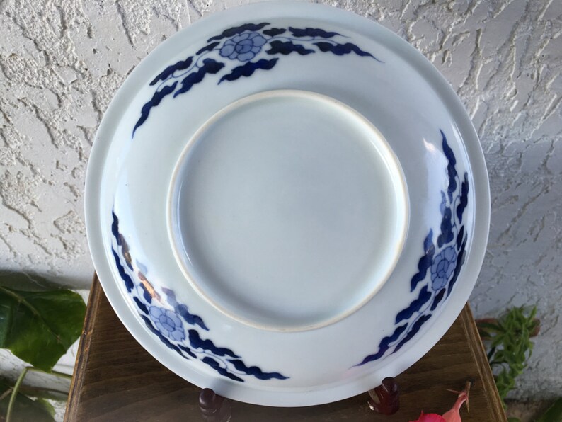 Op de afbeelding: Een witte keramische plaat met een blauw bloemen- en wolkenontwerp rond de rand. De plaat heeft een licht verhoogde rand en een gladde, glanzende afwerking. Het ontwerp is een traditioneel Aziatisch ge&iuml;nspireerd patroon.