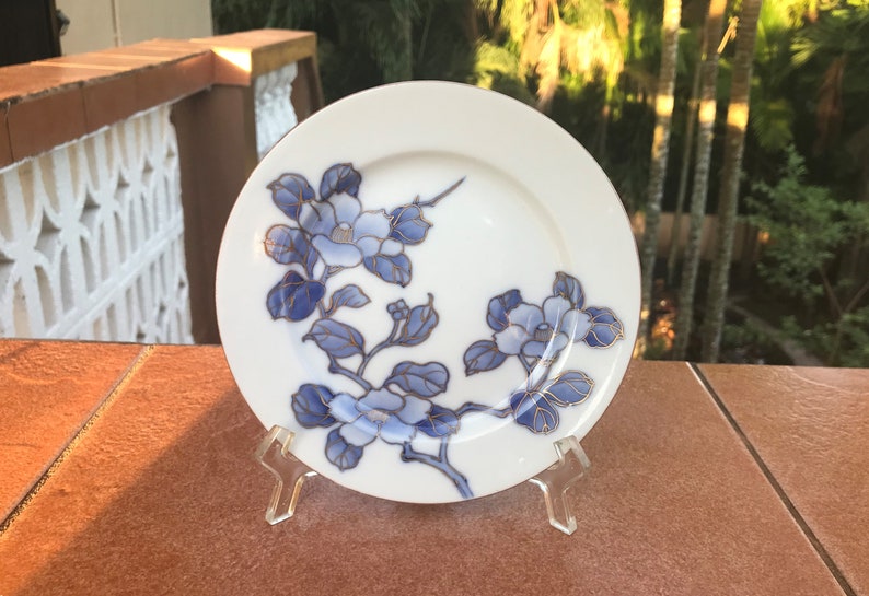 May include: White ceramic plate with a floral design in shades of blue and gold. The plate features a branch with blue flowers and leaves. The plate is displayed on a clear plastic stand, showcasing the decorative tableware.