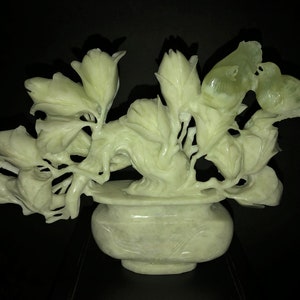 K&ouml;nnte beinhalten: Eine hellgr&uuml;ne, geschnitzte Steinskulptur von Blumen und V&ouml;geln in einer Vase. Die Skulptur zeigt detaillierte Bl&auml;tter, Bl&uuml;tenknospen und zwei V&ouml;gel, die auf &Auml;sten sitzen. Die Vase hat eine runde Form und steht auf einer dunklen Oberfl&auml;che.