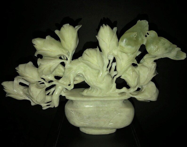 K&ouml;nnte beinhalten: Eine hellgr&uuml;ne, geschnitzte Steinskulptur von Blumen und V&ouml;geln in einer Vase. Die Skulptur zeigt detaillierte Bl&auml;tter, Bl&uuml;tenknospen und zwei V&ouml;gel, die auf &Auml;sten sitzen. Die Vase hat eine runde Form und steht auf einer dunklen Oberfl&auml;che.