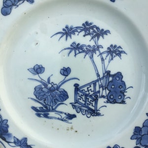 Op de afbeelding: Een wit porseleinen bord met een blauw en wit ontwerp van een tuinscene. Het ontwerp toont een bamboescherm, palmbomen en lotusbloemen.