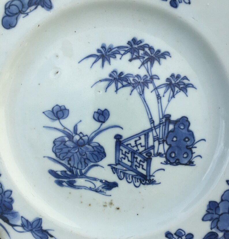 Op de afbeelding: Een wit porseleinen bord met een blauw en wit ontwerp van een tuinscene. Het ontwerp toont een bamboescherm, palmbomen en lotusbloemen.