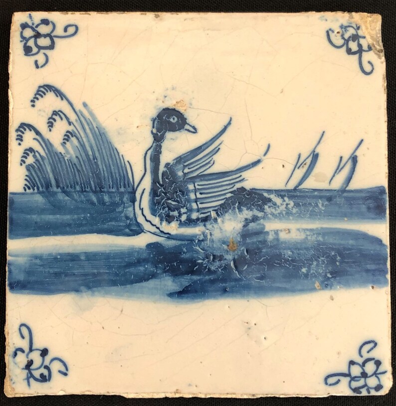 Puede incluir: Azulejo cer&aacute;mico cuadrado con un dise&ntilde;o pintado a mano de un pato nadando en agua azul. El pato est&aacute; representado con las alas extendidas y el agua tiene detalles de olas. Los acentos florales de las esquinas completan el dise&ntilde;o.
