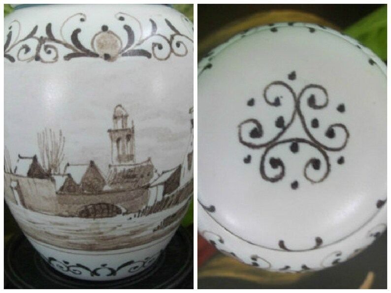 Puede incluir: Jarr&oacute;n de cer&aacute;mica blanca con una escena de paisaje en tonos sepia que representa un pueblo con una iglesia y un puente. El jarr&oacute;n tiene un borde y una parte superior decorativos con motivos florales marrones. La tapa tiene un dise&ntilde;o a juego.