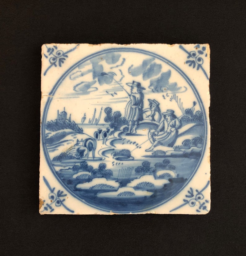May include: Square ceramic tile featuring a hand-painted blue and white pastoral scene. The scene shows figures fishing and herding animals near water, with trees and buildings in the background. Decorative floral corner accents.