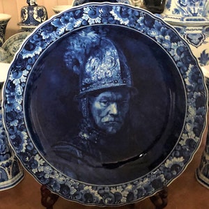 May include: A blue and white Delftware plate featuring a portrait of a man wearing a helmet. The plate is decorated with a floral border.