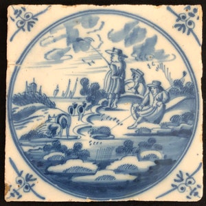 May include: A square, antique ceramic tile featuring a hand-painted pastoral scene in blue on a white background. The scene shows figures in hats, a river, trees, and a distant village. The tile is framed by a blue circular border and decorative corner elements.