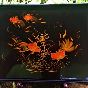 K&ouml;nnte beinhalten: Rechteckiges schwarzes Serviertablett mit handgemaltem Design von orangefarbenen Goldfischen und Wasserpflanzen. Das Tablett hat dekorative silberfarbene Kanten. Das Tablett wird von einem dunkelbraunen St&auml;nder getragen.