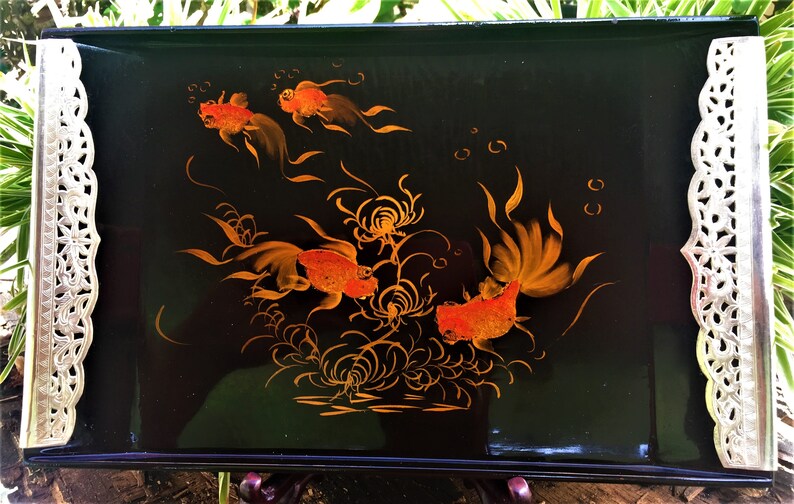 K&ouml;nnte beinhalten: Rechteckiges schwarzes Serviertablett mit handgemaltem Design von orangefarbenen Goldfischen und Wasserpflanzen. Das Tablett hat dekorative silberfarbene Kanten. Das Tablett wird von einem dunkelbraunen St&auml;nder getragen.