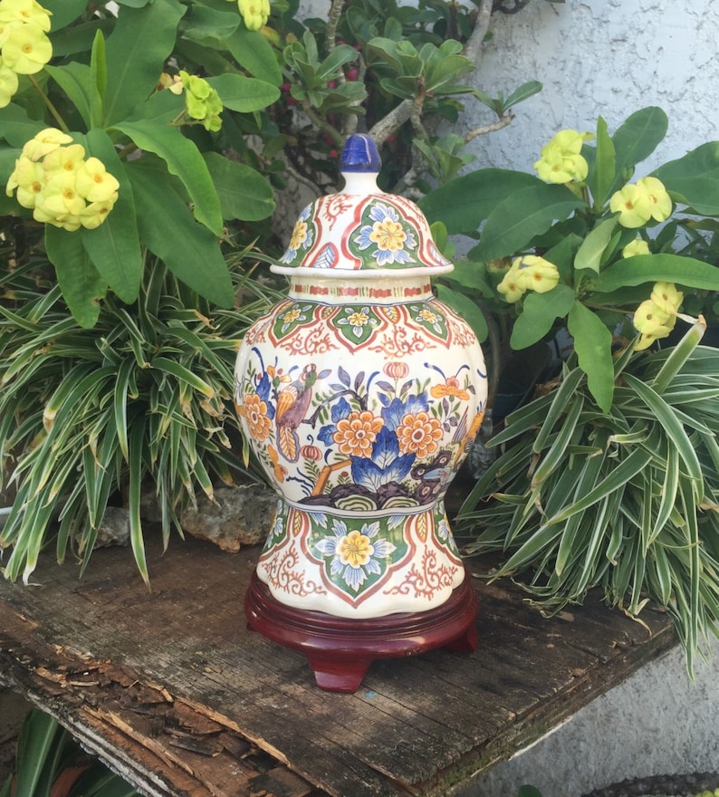 May include: A decorative ceramic ginger jar with a lid, featuring a colorful floral and bird design. The jar is set on a dark wooden stand. The color palette includes blue, orange, green, and white.
