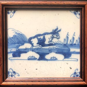 May include: A framed ceramic tile with a blue and white illustration of a rabbit or hare on a rock. The artwork depicts water and plants. The tile is set in a dark brown wooden frame, typical of traditional Delftware.