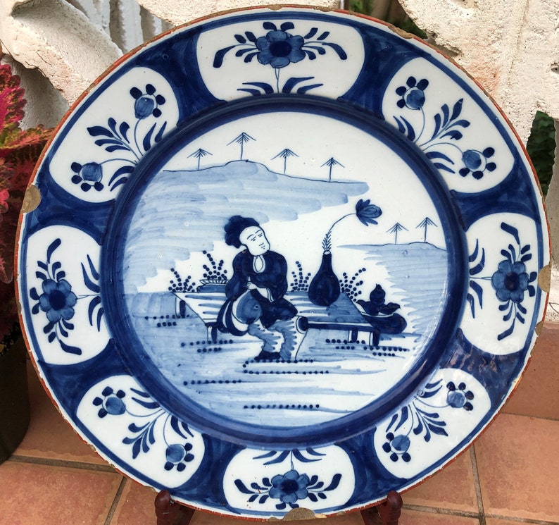 Puede incluir: Un plato de cer&aacute;mica azul y blanco con un borde decorativo y una escena de una persona sentada en un banco con un jarr&oacute;n de flores. El plato es de estilo Delft.