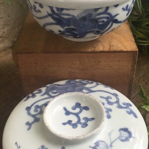 May include: White ceramic bowl and lid with blue floral and bird designs. The bowl is round with a matching lid and a small, circular knob. The lid features a detailed floral pattern. The bowl is sitting on a wooden surface.