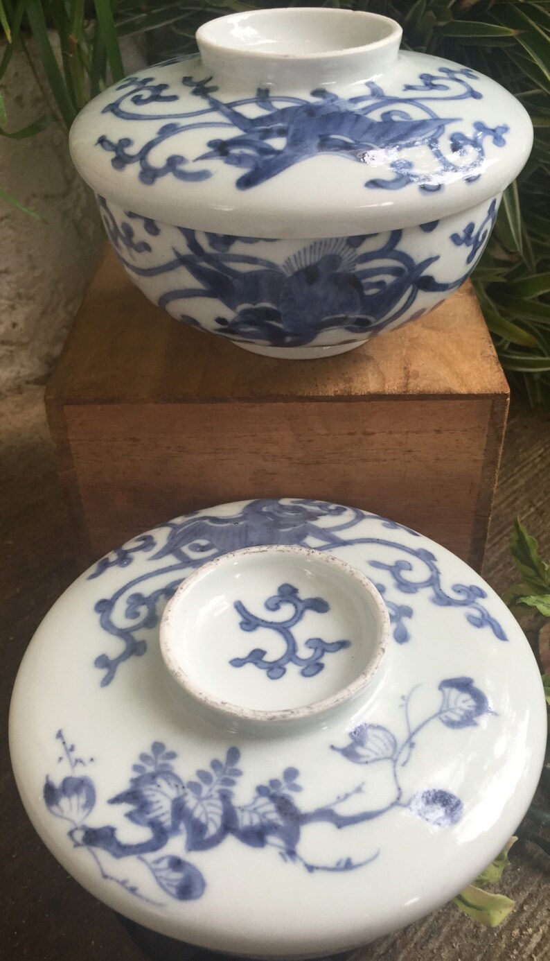 May include: White ceramic bowl and lid with blue floral and bird designs. The bowl is round with a matching lid and a small, circular knob. The lid features a detailed floral pattern. The bowl is sitting on a wooden surface.
