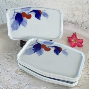 May include: White rectangular ceramic serving dishes with a blue and orange fruit and leaf design. The dishes are stacked, with one dish on top. A pink flower is in the background.