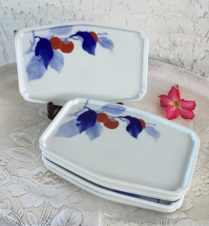 May include: White rectangular ceramic serving dishes with a blue and orange fruit and leaf design. The dishes are stacked, with one dish on top. A pink flower is in the background.
