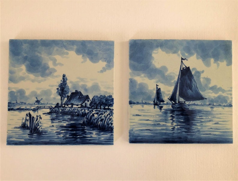 Puede incluir: Dos baldosas de cer&aacute;mica cuadradas con pinturas de paisajes azules y blancas. Una baldosa representa una escena rural con una casa, &aacute;rboles y un molino de viento. La otra muestra veleros en el agua bajo un cielo nublado.