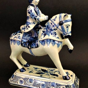 K&ouml;nnte beinhalten: Keramikfigur eines Reiters auf einem steigenden Pferd. Reiter und Pferd sind wei&szlig; mit blauen Blumenmustern. Der rechteckige Sockel hat ein passendes blaues Blumendesign. Der Reiter tr&auml;gt einen Hut und eine Jacke mit floralen Details.