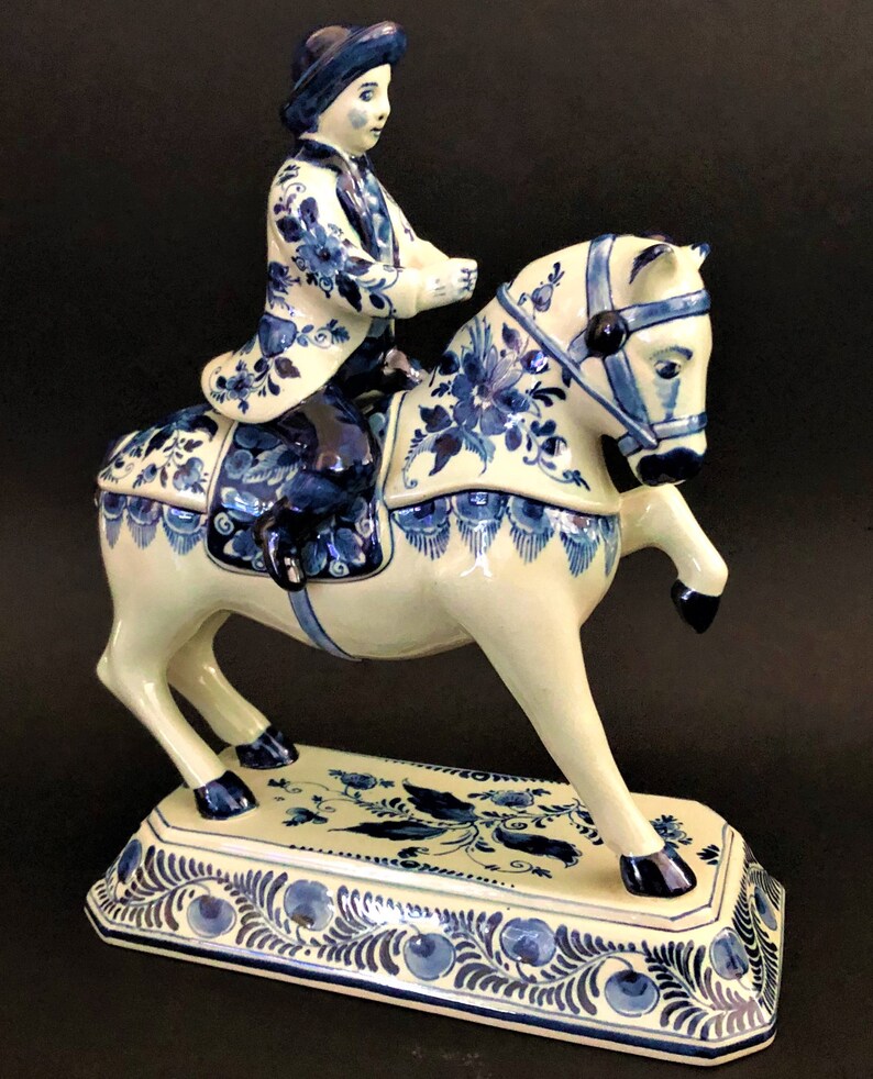 K&ouml;nnte beinhalten: Keramikfigur eines Reiters auf einem steigenden Pferd. Reiter und Pferd sind wei&szlig; mit blauen Blumenmustern. Der rechteckige Sockel hat ein passendes blaues Blumendesign. Der Reiter tr&auml;gt einen Hut und eine Jacke mit floralen Details.