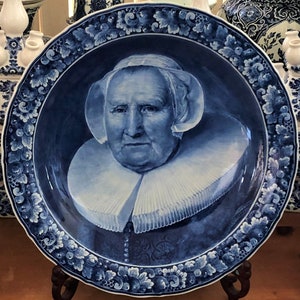May include: A blue and white Delftware plate featuring a portrait of a woman wearing a white lace collar and a white cap. The plate is decorated with a floral border.