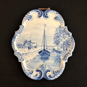 May include: A blue and white ceramic wall plaque featuring a winter scene with a sailboat on a canal, a house, and trees. The plaque has a decorative, ornate border. A brown string is attached for hanging.