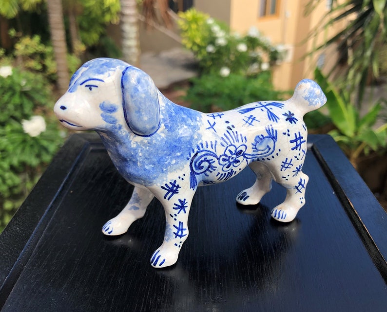 Puede incluir: Figura de perro de cer&aacute;mica en blanco y azul. El perro tiene orejas azules y una cabeza salpicada de azul. El cuerpo es blanco con patrones florales y geom&eacute;tricos azules. La figura est&aacute; de pie sobre cuatro patas con una peque&ntilde;a cola.