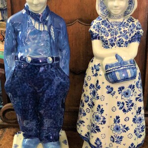 K&ouml;nnte beinhalten: Zwei Keramikfiguren, die ein holl&auml;ndisches Paar in traditioneller Kleidung darstellen. Der Mann tr&auml;gt eine blaue Uniform und Hut, die Frau ein Blumenkleid und eine Haube. Beide sind blau-wei&szlig; bemalt und stehen auf dekorativen Sockeln.