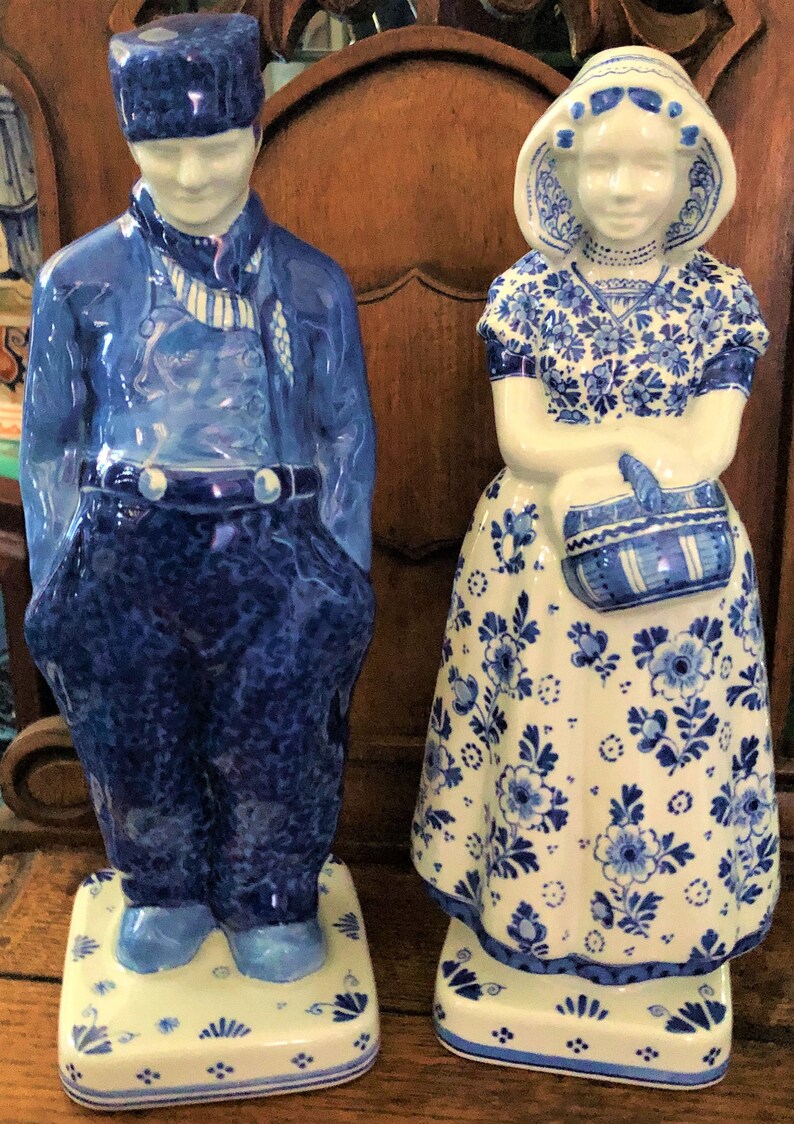 K&ouml;nnte beinhalten: Zwei Keramikfiguren, die ein holl&auml;ndisches Paar in traditioneller Kleidung darstellen. Der Mann tr&auml;gt eine blaue Uniform und Hut, die Frau ein Blumenkleid und eine Haube. Beide sind blau-wei&szlig; bemalt und stehen auf dekorativen Sockeln.