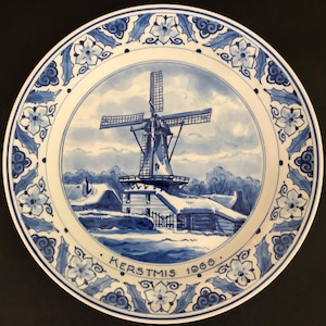 May include: Blue and white Delftware plate featuring a winter scene of a windmill and houses. The plate has a blue and white floral border and the text "KERSTMIS 1966".