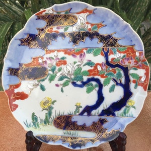 May include: A decorative, scalloped-edge plate with a Japanese-inspired design. The plate features a blue border, a central scene with a blue tree, white flowers, and accents of gold, red, and yellow. The plate is displayed on a wooden stand.