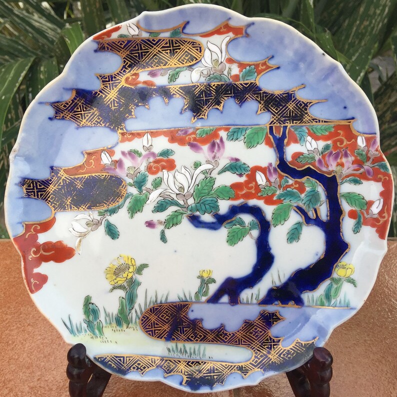 May include: A decorative, scalloped-edge plate with a Japanese-inspired design. The plate features a blue border, a central scene with a blue tree, white flowers, and accents of gold, red, and yellow. The plate is displayed on a wooden stand.