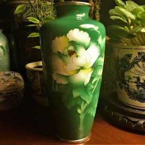 Puede incluir: Un jarr&oacute;n de cer&aacute;mica verde con un dise&ntilde;o floral blanco. El jarr&oacute;n tiene un borde plateado y un acabado brillante.