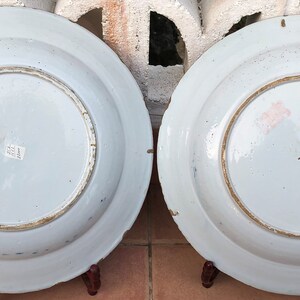 Puede incluir: Dos platos grandes de cer&aacute;mica blanca con dise&ntilde;os pintados en azul y oro. Los platos est&aacute;n expuestos en soportes de madera.