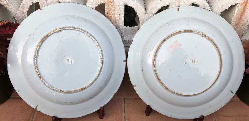 Puede incluir: Dos platos grandes de cer&aacute;mica blanca con dise&ntilde;os pintados en azul y oro. Los platos est&aacute;n expuestos en soportes de madera.