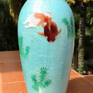 Pu&ograve; includere: Un vaso in ceramica blu turchese con un pesce dorato e piante verdi dipinte sul lato. Il vaso ha una superficie testurizzata e si trova su una base in legno nero.