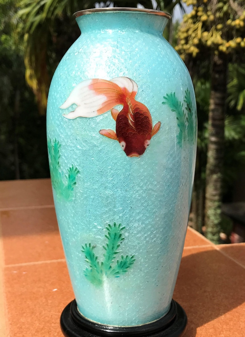 Pu&ograve; includere: Un vaso in ceramica blu turchese con un pesce dorato e piante verdi dipinte sul lato. Il vaso ha una superficie testurizzata e si trova su una base in legno nero.