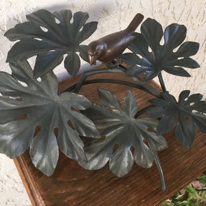 Puede incluir: Una escultura de p&aacute;jaro de bronce posada en una rama de metal con hojas grandes y estilizadas. Las hojas son de un color verde gris&aacute;ceo apagado. La escultura est&aacute; montada sobre una superficie de madera, lo que sugiere un art&iacute;culo decorativo para el hogar.