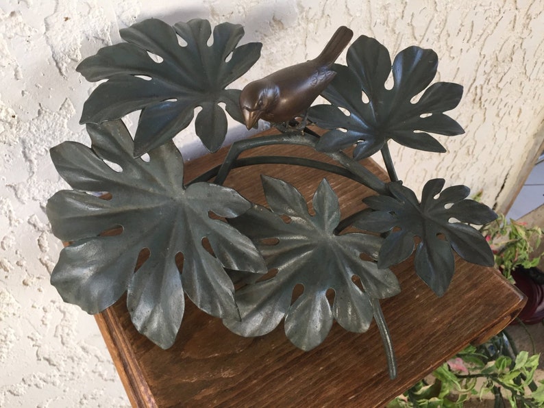Puede incluir: Una escultura de p&aacute;jaro de bronce posada en una rama de metal con hojas grandes y estilizadas. Las hojas son de un color verde gris&aacute;ceo apagado. La escultura est&aacute; montada sobre una superficie de madera, lo que sugiere un art&iacute;culo decorativo para el hogar.