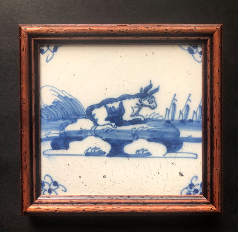 Puede incluir: Azulejo de cer&aacute;mica enmarcado cuadrado con una ilustraci&oacute;n azul y blanca de una vaca o un toro descansando sobre una roca. La obra de arte est&aacute; enmarcada en un marco de madera marr&oacute;n oscuro. El azulejo tiene una est&eacute;tica tradicional y antigua.