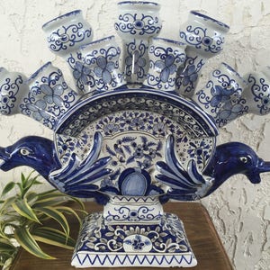 May include: A white and blue ceramic tulip vase with a dolphin design. The vase has multiple tulip holders, a decorative plate, and a pedestal base. The blue floral and swirl patterns add a classic touch to this decorative piece.