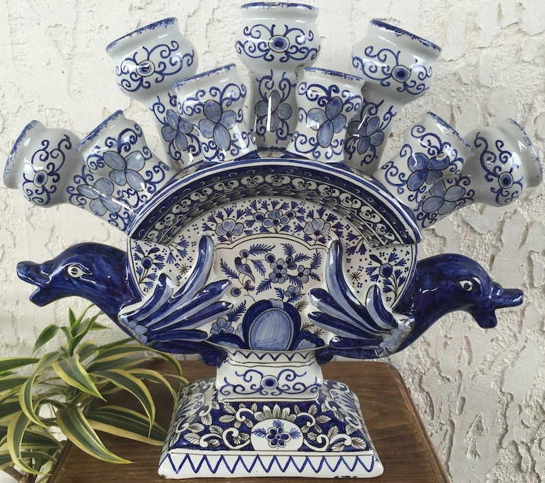 May include: A white and blue ceramic tulip vase with a dolphin design. The vase has multiple tulip holders, a decorative plate, and a pedestal base. The blue floral and swirl patterns add a classic touch to this decorative piece.