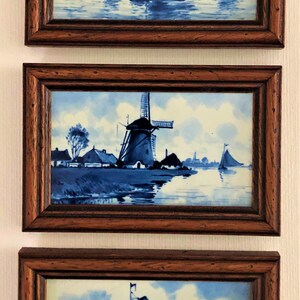 Puede incluir: Tres pinturas enmarcadas al estilo Delft. Cada una presenta una escena n&aacute;utica azul y blanca con veleros y molinos de viento. Los marcos son de madera oscura. Las pinturas se exhiben verticalmente en una pared blanca.