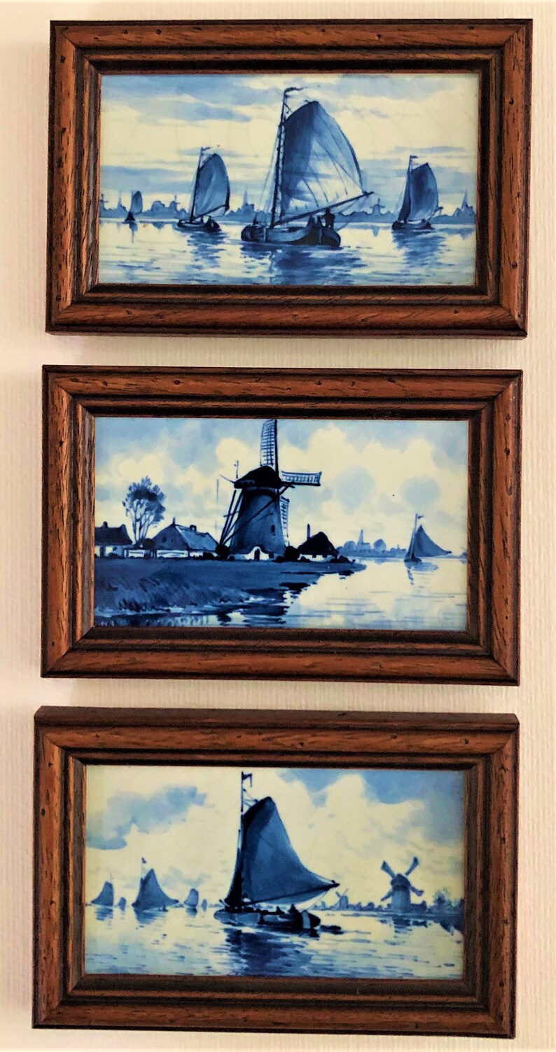 Puede incluir: Tres pinturas enmarcadas al estilo Delft. Cada una presenta una escena n&aacute;utica azul y blanca con veleros y molinos de viento. Los marcos son de madera oscura. Las pinturas se exhiben verticalmente en una pared blanca.