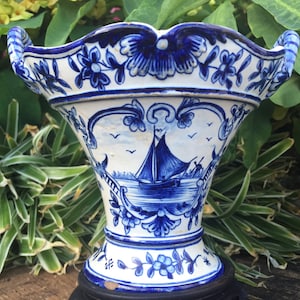 May include: A white ceramic vase featuring blue hand-painted illustrations of a sailboat on water, surrounded by floral motifs and scrollwork. The vase has a flared, scalloped rim with two side handles and sits on a dark circular base.