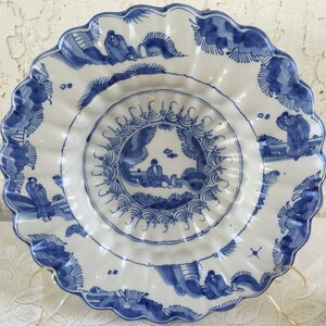 Puede incluir: Un plato grande de cer&aacute;mica blanca con un dise&ntilde;o azul y blanco. El plato tiene un borde festoneado y presenta un dise&ntilde;o central de una figura en un paisaje. El plato est&aacute; decorado con &aacute;rboles y flores estilizados.