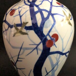 May include: A white porcelain vase with a blue and red floral design. The vase has a gold rim and features a stylized tree with birds and red berries.