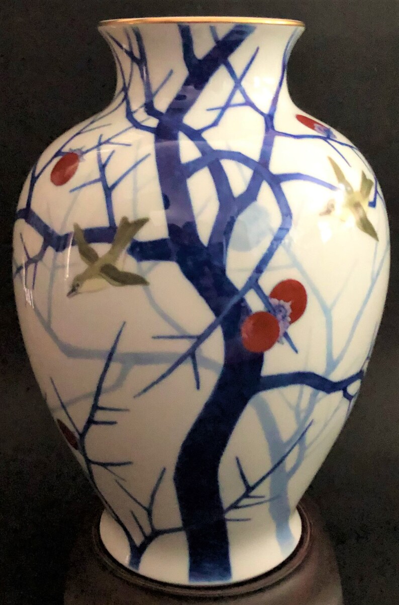 May include: A white porcelain vase with a blue and red floral design. The vase has a gold rim and features a stylized tree with birds and red berries.